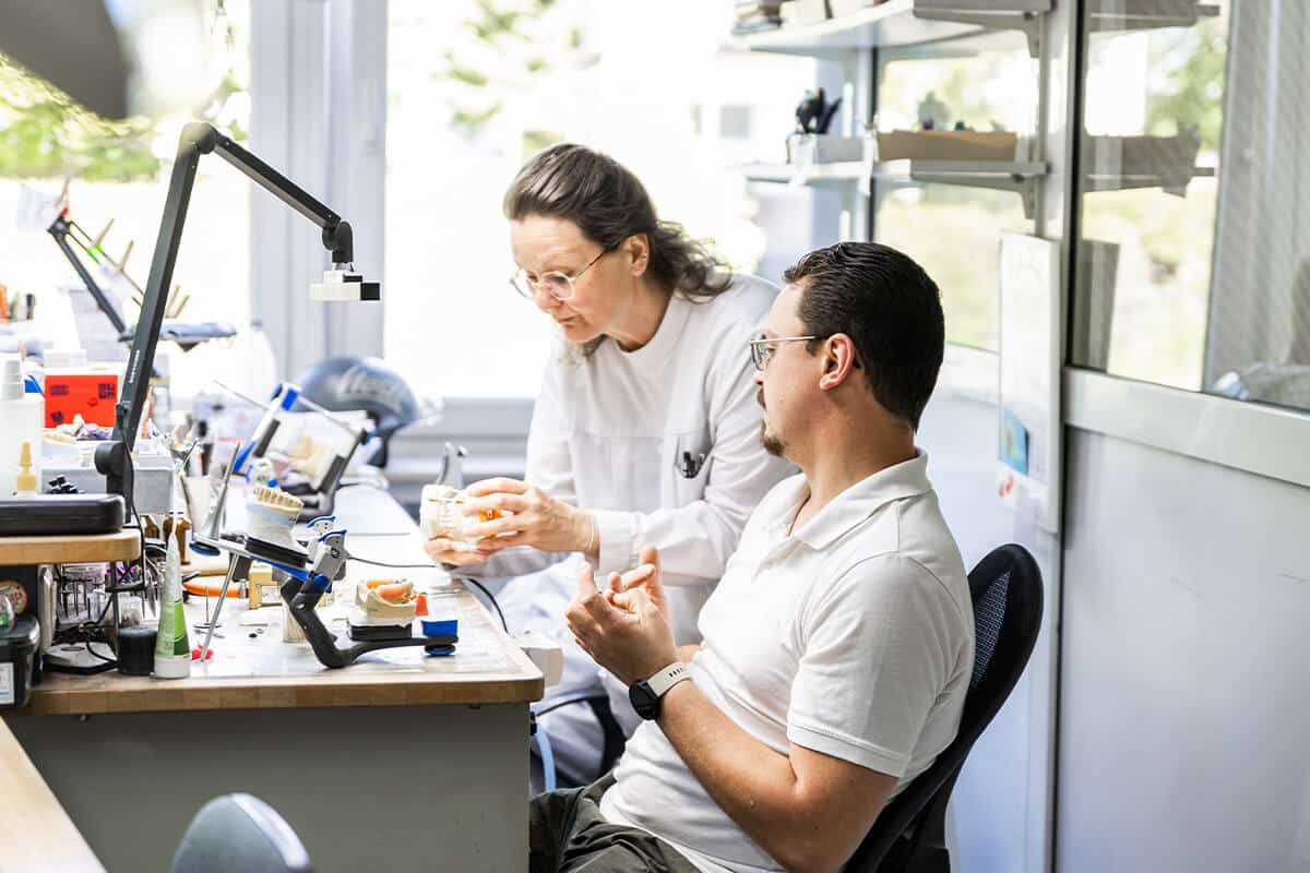 Zwei Personen in weißer Arbeitskleidung sitzen an einem Tisch mit technischen Geräten und Werkzeugen.