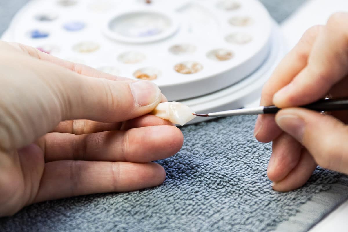 Hände halten und bemalen eine kleine Zahnprothese mit einem feinen Pinsel vor einer Farbpalette.