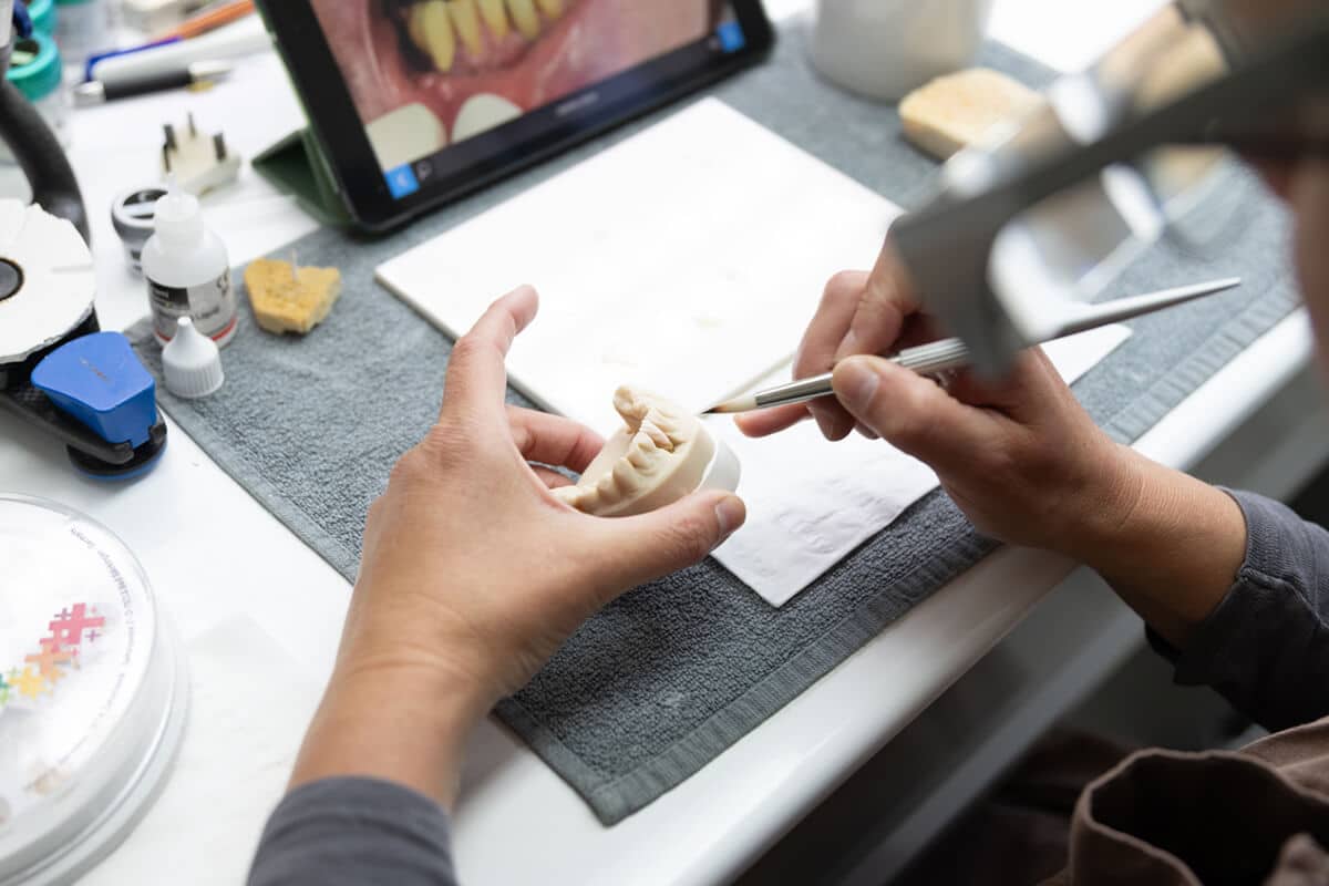 Hände halten und bearbeiten ein Zahnmodell mit einem Pinsel, im Hintergrund ein Tablet mit Zahnbildern.