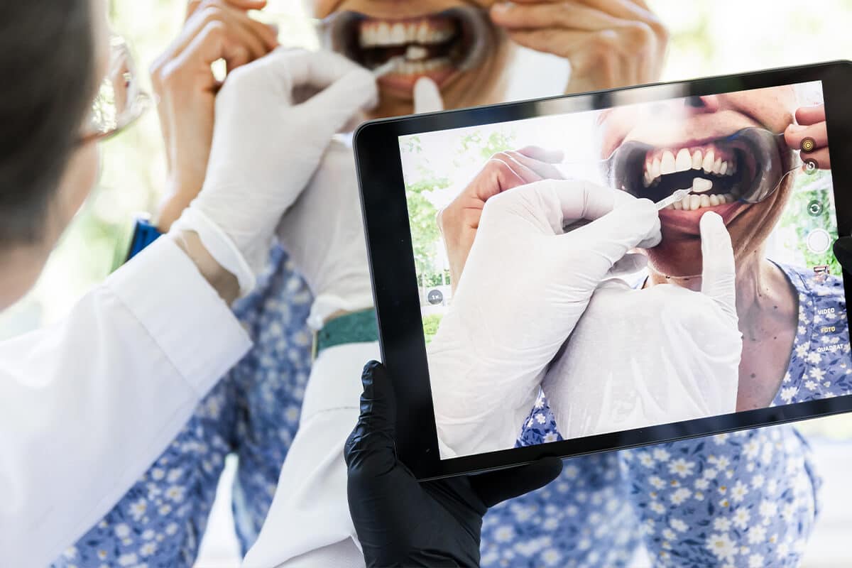 Person hält Tablet mit Nahaufnahme eines geöffneten Mundes, während eine andere Person mit Handschuhen die Zahnfarbe abstimmt.