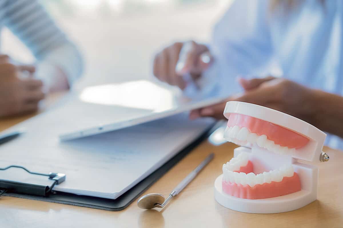 Person zeigt Patientin auf einem Tablet etwas, im Vordergrund liegt ein Zahnmodell und ein zahnärztliches Instrument auf dem Tisch.